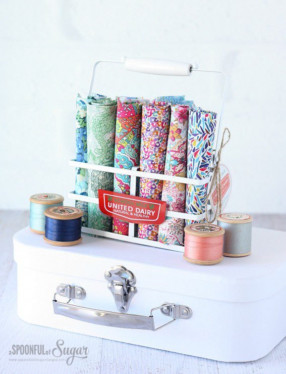 Bundle of Liberty Tana Lawn fabric in a basket sitting on top of a white mini suitcase. Wooden cotton reels with brightly coloured thread sitting next to the fabric.