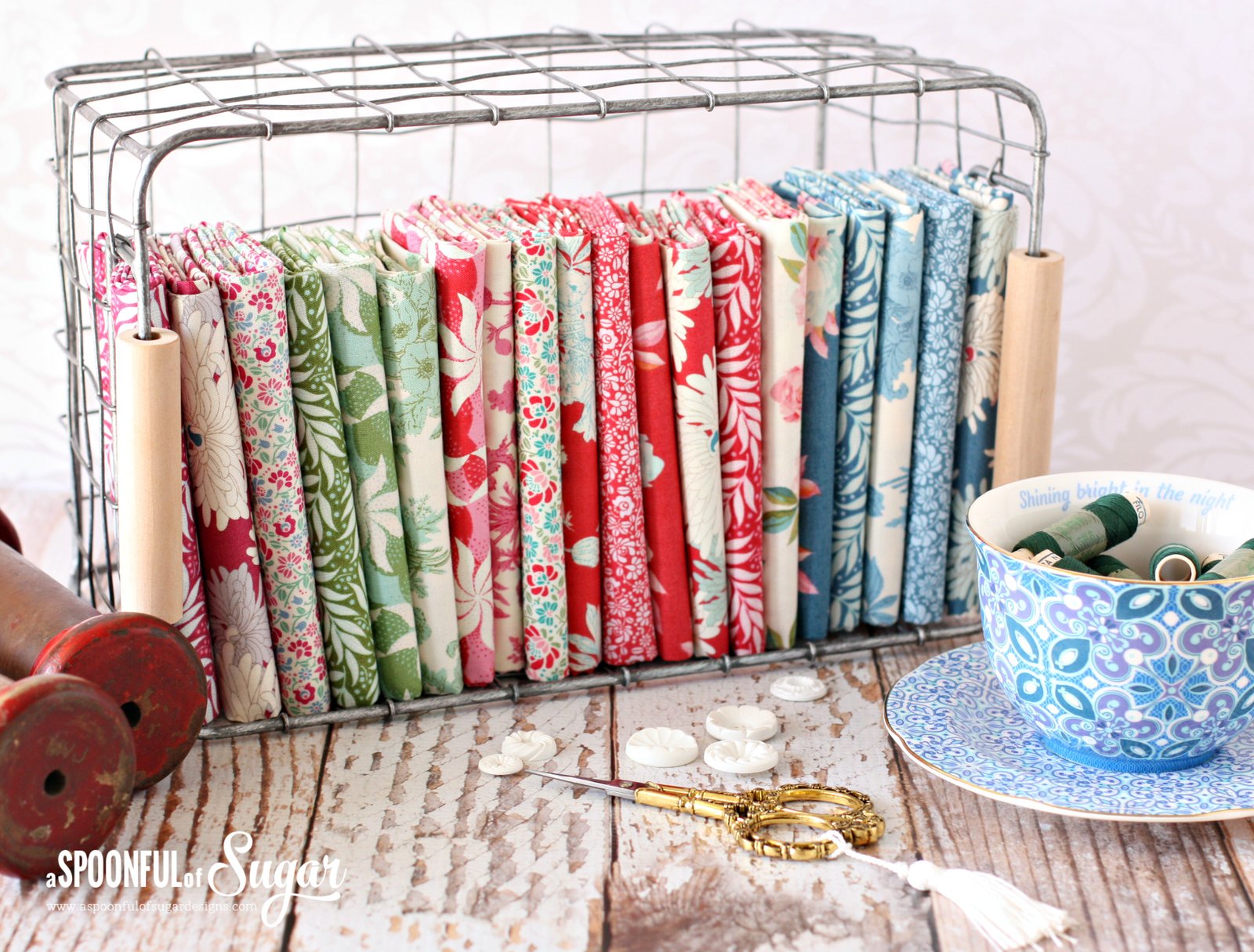 basket of lovely tilda fabrics in shades of green, red, and blue. Tea cup, buttons and scissors in foreground.