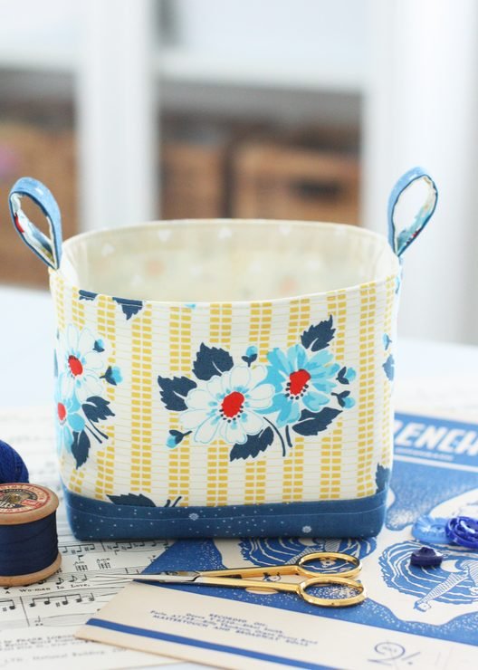 A Luma Bucket (fabric basket) sitting on a table with scissors and thread and buttons next to it.