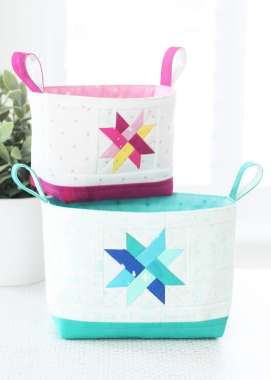 Two Luma Bucket fabric organisers in bright Ruby Star Society prints, one pink and one green, shown side‑by‑side to display the woven star panel and contrast base. Pattern by A Spoonful of Sugar Designs. 