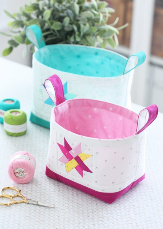 Two Luma Bucket fabric organisers in bright Ruby Star Society prints, one pink and one green, shown side‑by‑side to display the woven star panel and contrast base. Pattern by A Spoonful of Sugar Designs. 