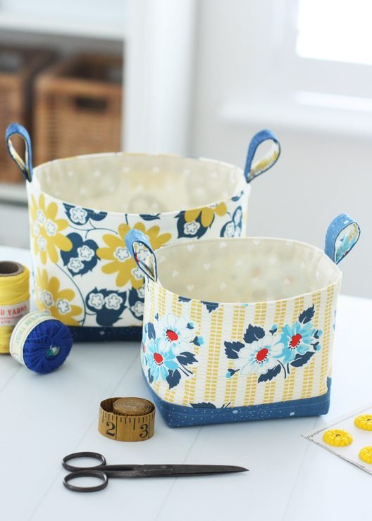Pair of Luma Fabric Buckets made in navy, mustard, and red fabric. Sitting on a table with scissors, tape measure and thread next to it.